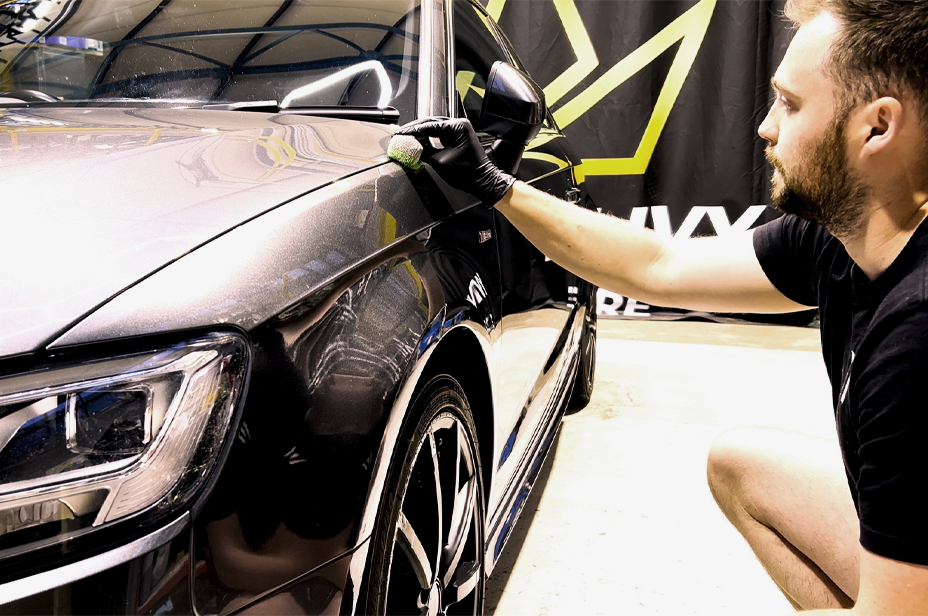 Person applying car wax to a vehicle with a brand flag in the background