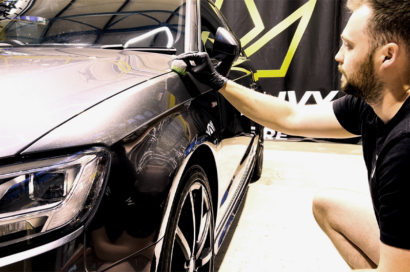 Person applying car wax to a vehicle with a brand flag in the background